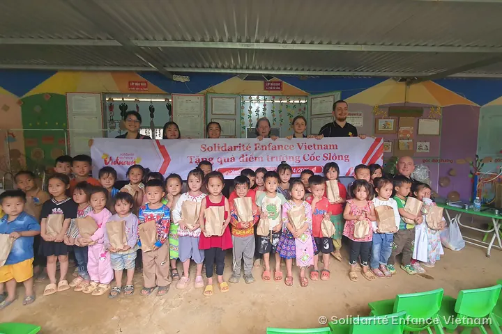 Des enfants de maternelle, un petit sac en papier dans les mains, posent devant la banderole de l’association à l’école de Cốc Slông.