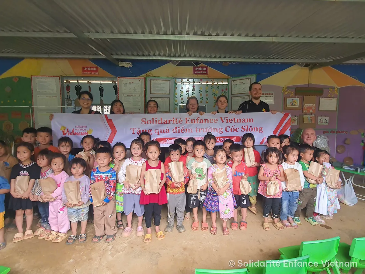 Des enfants de maternelle, un petit sac en papier dans les mains, posent devant la banderole de l’association à l’école de Cốc Slông.