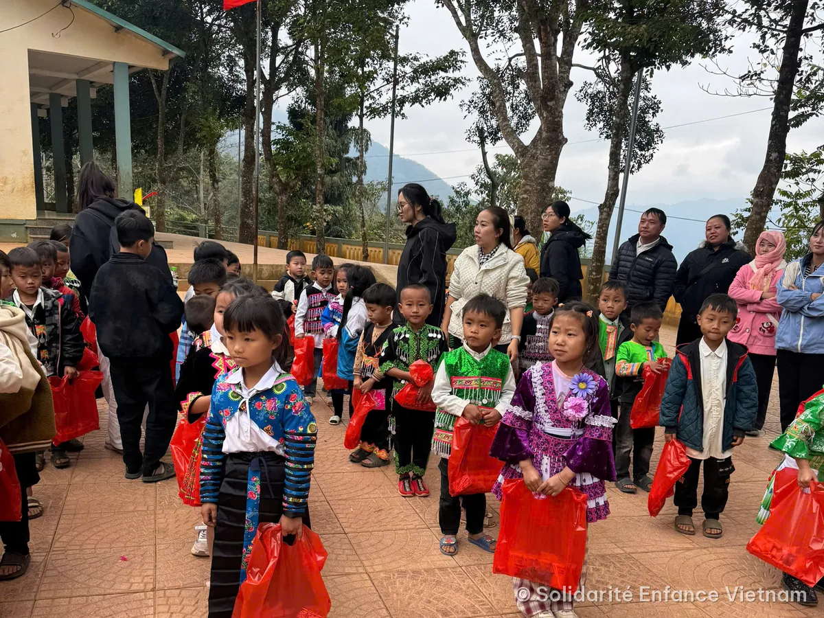 Des enfants en costumes traditionnels, chacun avec un sac rouge, dans la cour de l’école.