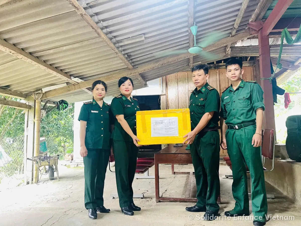 Remise d’un carton de médicaments à des personnes en uniforme vert, sous un auvent.